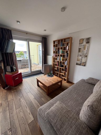 Heller Wohnraum mit Sofa, Couchtisch, TV und Bücherregal, Blick auf den Balkon.